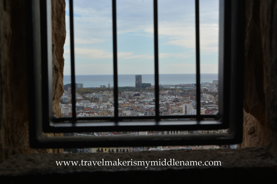 A metal grate covers openings cut into the stone walls of the Passion tower in La Sagrada Familia church in Barcelona, Spain, and shows a view of the Meditteranean sea in the distance.