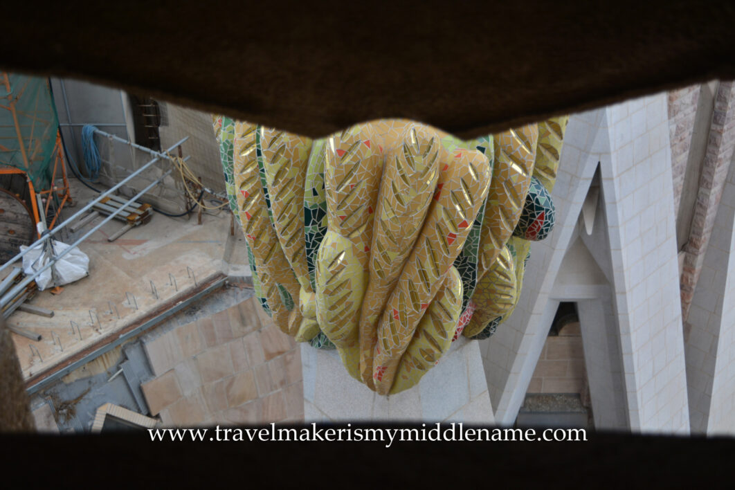 A close up look of a yellow fruit sculpture that looks like twisted finger limes atop a peak at La Sagrada Familia church in Barcelona, Spain, as seen through a small cutout in the stone wall.