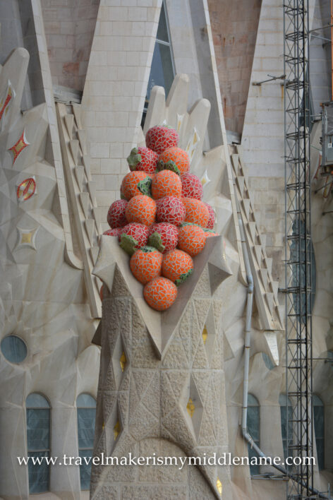 A sculpture of a cluster of red-orange balls representing fruit in La Sagrada Familia church in Barcelona, Spain. White triangular stone structures are in the background. 
