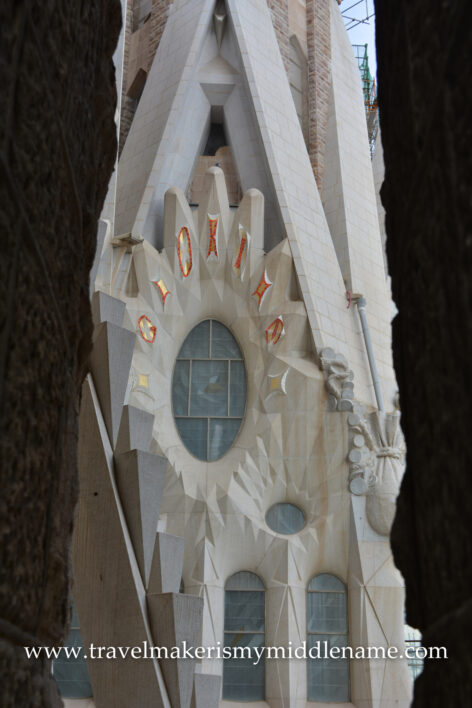 Detail of the facade of La Sagrada Famiila church in Barcelona, Spain, at the tower height. A portrait oriented oval glass window has spikey stoney rays emanating from the glass, like a vertically oriented eye.