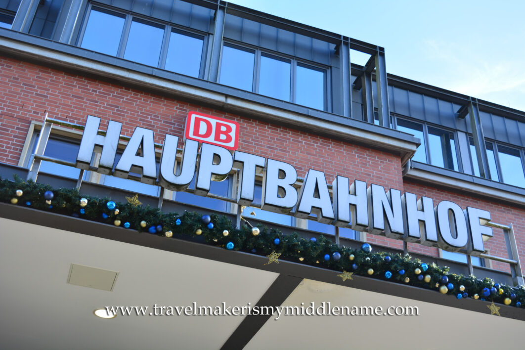 A sign saying "Hauptbahnof" outside a central train station in Germany with Christmas lights underneath, against a blue sky.