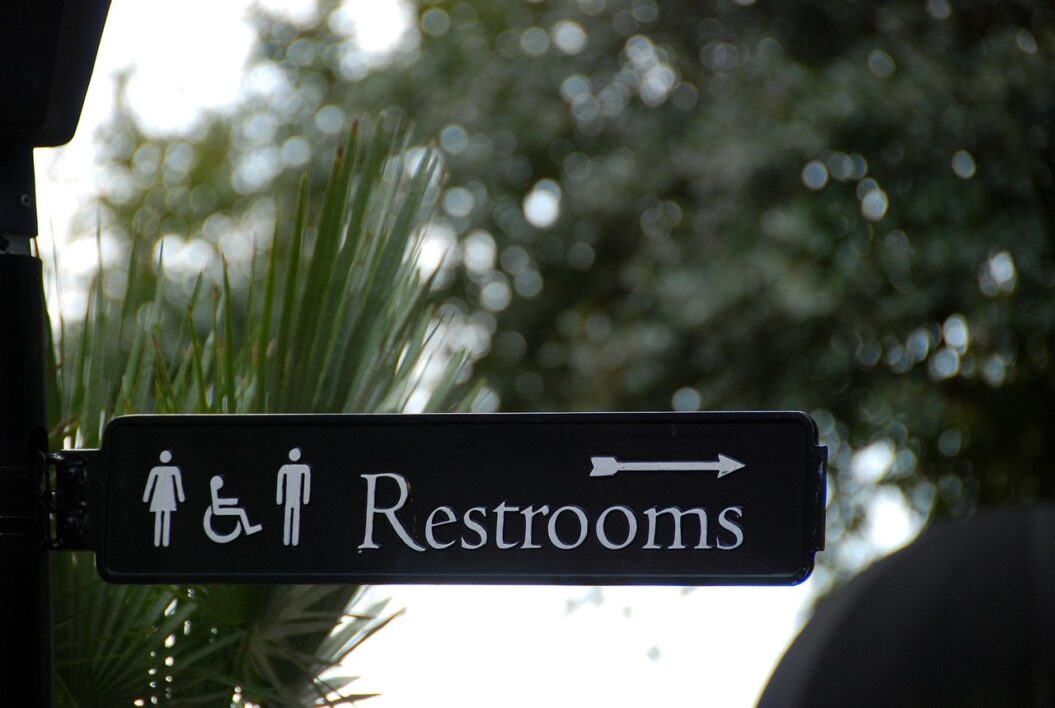A sign with a white woman, a wheelchair, and a man icon followed by the word "Restrooms" and an arrow pointing to the right, outside, day, with a tree canopy in the background.